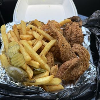 Chick N Headz - A little bit of everything was good! - The image shows a takeout meal with fried chicken pieces, French fries, and pickles in a white foam container lined with aluminum foil. The fried chicken includes a drumstick and other pieces with crispy, seasoned coating. The French fries are golden and slightly seasoned, and there are sliced pickles on the side. The container is placed inside a black plastic bag, with a white foam box in the background, suggesting a casual food delivery or takeout setting. - Food Trucks Near Me - Forney, Texas
