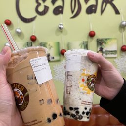 Coffee Frap - Two individuals display cups of bubble tea, one with brown tapioca pearls and the other with a cream-colored drink, in front of a tea shop wall decorated with ornaments and artwork.