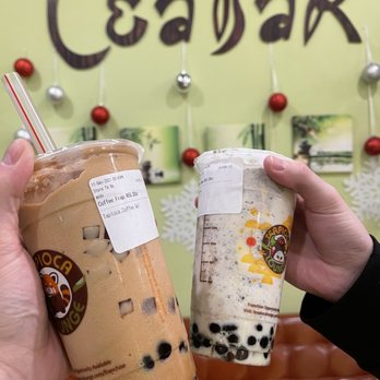 Teapioca Lounge - Coffee Frap - Two individuals display cups of bubble tea, one with brown tapioca pearls and the other with a cream-colored drink, in front of a tea shop wall decorated with ornaments and artwork. - Bubble Tea Near Me - Lubbock, Texas