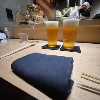 Hiden - The image shows two glasses of beer with foam on top, placed on black coasters on a light wooden counter at a sushi restaurant. A folded dark cloth napkin, a pair of chopsticks, and a small water glass are also on the counter. In the background, a chef wearing a white uniform and face mask is working in the kitchen area, which features stainless steel appliances and a wooden wall. The setting appears to be a modern, minimalist sushi bar or omakase restaurant. - Japanese Near Me - Miami, Florida