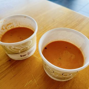 Toss’d - Tomato Bisque soup of the day comes with entrees from 11-2pm - Two disposable cups filled with tomato bisque soup are positioned on a wooden table. The cups have a decorative design and contain a reddish-orange soup with visible small black specks. The cups are partially used, with some soup residue on the inner rims. The background shows a tiled floor, indicating an indoor setting, likely a restaurant or cafe. - Wraps Near Me - Temecula, California