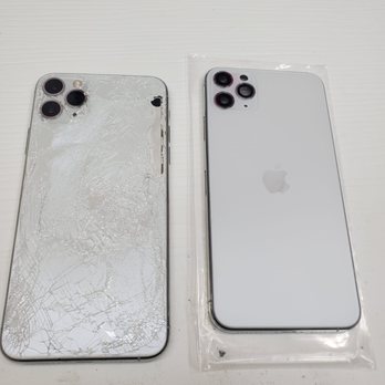 Ucrackifix - Cambio de back glass iPhone 11 Pro Max muy excelente trabajo - The image features two iPhones on a white surface. The phone on the left has a severely cracked back glass with visible shattering and damage, while the phone on the right is intact, with a clean, undamaged back and is partially covered in a clear plastic sleeve. Both phones have triple-camera setups in the top left corner. The setting appears to be a repair shop or similar environment. - Mobile Phone Repair Near Me - Trenton, New Jersey