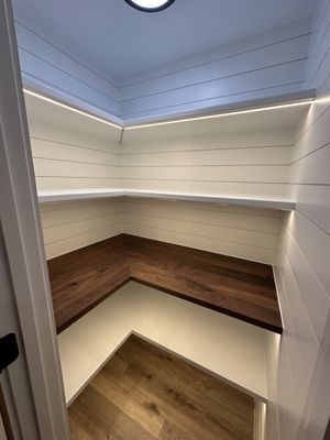 Pantry Upgrade - This pantry remodel showcases a compact, L-shaped layout with clean white shiplap walls for a modern farmhouse vibe. Floating white shelves line the upper walls, each equipped with subtle LED strip lighting underneath for even illumination. A custom walnut countertop in a rich, dark finish serves as the centerpiece, providing a warm, durable surface for prep or storage, while lower shelves and hardwood flooring complete the functional, inviting space.