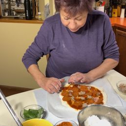 A woman with short, curly hair is preparing a pizza in a home kitchen. She is placing toppings on a dough base covered with tomato sauce. The countertop has bowls of ingredients including cheese, meat, and vegetables, along with kitchen utensils and appliances in the background.