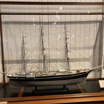 Marine Museum of Fall River - A detailed scale model of a sailing ship with three masts and intricate rigging is showcased inside a glass display case at a museum. The ship features a black hull with white accents and is mounted on a black base. The background includes museum walls and additional exhibits, with reflections of lights visible on the glass surface. An informational plaque is positioned on the right side of the display. - Museums Near Me - Fall River, Massachusetts