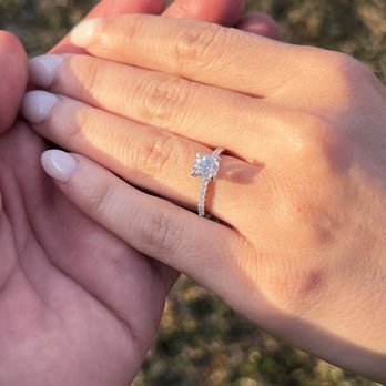 Atlantic Gems - Custom Diamond Engagement Ring - The image depicts a hand with a diamond engagement ring featuring a large, round-cut central diamond and a thin band encrusted with smaller diamonds. The hand is positioned against a blurred outdoor background, possibly grass or ground, with warm lighting highlighting the ring and skin tones. The focus is on the ring and the hand, emphasizing the jewelry detail. - Jewelry Near Me - Silver Spring, Maryland