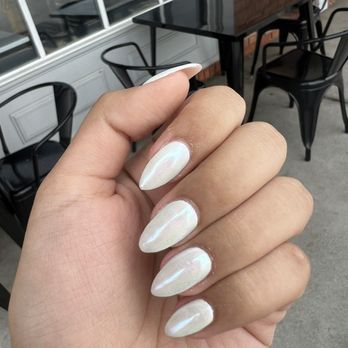 Style Nails - The image shows a hand with five fingers, each with long, almond-shaped nails painted in a shiny, white, iridescent polish. The hand is positioned in front of a cafe or outdoor seating area with black chairs and a dark wooden table, with a brick wall and window in the background. The setting appears to be outside a restaurant or cafe. - Nail Salons Near Me - Bridgewater, New Jersey