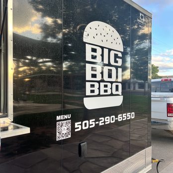 Big Boi BBQ - Food Truck - The image shows a black food truck with a large white logo of a burger and the text 'BIG BOI BBQ' on its side. The truck displays a QR code labeled 'MENU' and a phone number, 505-290-6550. It is parked outdoors, with a reflection of the sky and trees on its surface, and other vehicles are visible nearby. - Food Trucks Near Me