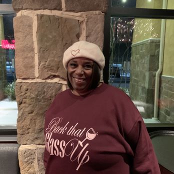The Cave - A woman in a white beret and maroon sweatshirt with the phrase "Buck that Glass Up" is standing inside a restaurant or cafe, with a stone wall and large window behind her. - Wine Tasting Room Near Me - Canton, Ohio