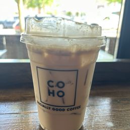 Iced Chai Latte - A plastic cup containing iced coffee with almond milk, topped with foam, is positioned on a wooden table near a window. The cup displays the CoHo Coffee House logo and slogan, and the background shows a bright outdoor scene with trees and parked cars. The image emphasizes the beverage's appearance and the coffee shop's branding.