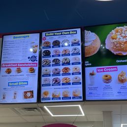 Two digital menu screens show various donuts, beverages, breakfast sandwiches, and ice cream options at a donut shop.
