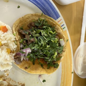Las Islitas Stone Park - A taco with beef, chopped cilantro, and diced onions is placed on a colorful, striped plate. The plate also contains a serving of rice with corn and other ingredients. A white cup and a straw are visible on the table, indicating a casual dining setting. - Mexican Near Me - Stone Park, Illinois