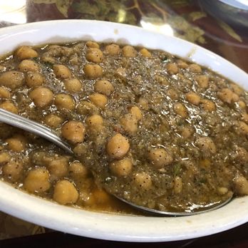 Redchilli - Chana Masala - A chickpea curry is presented in an oval white bowl, featuring cooked chickpeas in a thick, seasoned sauce with visible herbs and spices. A metal spoon rests inside the bowl, and part of a patterned tablecloth and a bowl are visible in the background. - Indian Near Me - Selden, New York