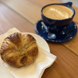 a croissant and a cup of coffee - A croissant on a white plate with a napkin and a cup of coffee in a blue cup and saucer on a wooden table.