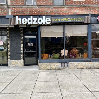Hedzole - The image shows the exterior of Hedzole, a Ghanaian-American restaurant, with a black facade, large glass window, and stone accents at the base. The sign above the entrance displays the name "hedzole" in large white letters and the phrase "PAN AFRICAN SOUL" in yellow. Inside, a few people are seated near the window, and the sidewalk in front is wide and clean. The building above has a brick exterior with several windows and air conditioning units. - African Near Me - Washington, DC, District of Columbia