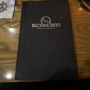 Buck & Honey's - A black textured menu cover with the Buck & Honey's restaurant logo is placed on a wooden table, with a glass of water and a napkin nearby. - American Near Me - Monona, Wisconsin
