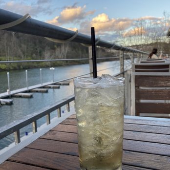 Table 9 - A tall, clear glass filled with a light-colored iced beverage and a black straw is positioned on a wooden table outdoors. The background features a river, a railing, and a sky with scattered clouds, suggesting a riverside setting during late afternoon or early evening. - New American Near Me - Morgantown, West Virginia