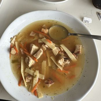 Offerdahl's Off-The-Grill - Chicken soup - A white bowl filled with chicken soup featuring shredded chicken pieces, julienned carrots, and other vegetables in a clear broth. A metal spoon rests on the edge of the bowl, partially submerged in the soup. The bowl is placed on a white table, with a plate of bread and some condiment packets visible in the background. - Sandwiches Near Me - Lighthouse Point, Florida