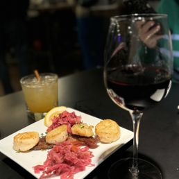 Scallops, cabernet and a funky drink! - A plate of seafood including seared scallops, fish, and pickled vegetables is presented on a black table. A glass of red wine is positioned in the foreground, partially obscuring the plate, while a cocktail with a straw is visible in the background. The setting appears to be a restaurant with a dark interior and other patrons in the background.