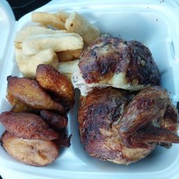1/2 White Chicken - A white takeout container holds a roasted chicken quarter, fried plantains, and yuca fries. The chicken has a crispy, browned skin, and the plantains are golden and fried. The yuca fries are light-colored and crispy. The container is partially open, revealing the food inside, with no visible sauces or additional garnishes.