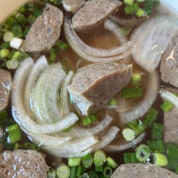 Pho Saigon - Meatball pho - A bowl of Vietnamese meatball pho contains sliced onions, chopped green onions, and meatballs submerged in clear broth. The ingredients are arranged with the onions and green onions floating on top, and the meatballs are visible throughout the bowl. The broth appears light and transparent, typical of traditional pho dishes. - Soup Near Me - Dover, Delaware