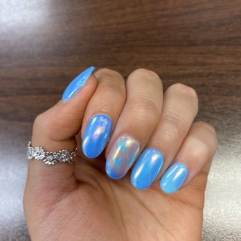 Luxury Nails - Overlay Natural Nail - A hand with five fingernails painted in a glossy blue color, with the middle nail featuring a holographic, iridescent design. The hand is wearing a silver ring with floral motifs on the ring finger. The background is a wooden surface, and the image focuses on the nail art and jewelry, showcasing a modern manicure style. - Nail Salons Near Me - Elmhurst, Illinois