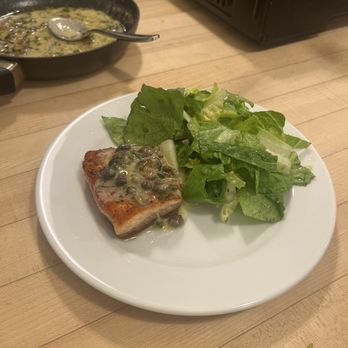 Sur La Table - A white round plate contains a cooked salmon fillet topped with a sauce that includes green herbs and capers. Next to the salmon is a side salad made of leafy greens, including romaine and possibly spinach. The plate is placed on a light-colored wooden table. In the background, there is a black skillet with remnants of sauce and a spoon, and a portable stove or grill is partially visible. - Kitchen Supplies Near Me - Glendale, Wisconsin