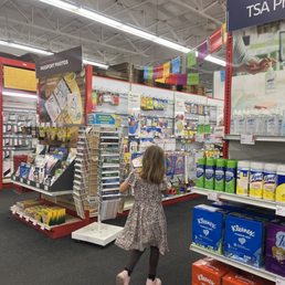 A young girl with shoulder-length hair, dressed in a patterned dress and dark leggings, is walking through an aisle in a retail store. She is facing away from the camera and appears to be looking at various office supplies, including notebooks, pens, and other stationery items displayed on shelves and racks. The store has bright lighting, a high ceiling with exposed beams, and colorful decorations hanging overhead.