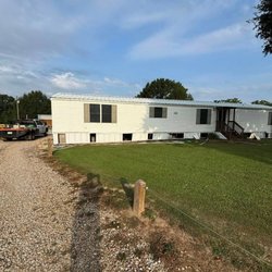 A wide exterior view of a white mobile home with a newly installed white metal roof and skirting, set on a green lawn.