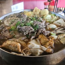 Taiwanese Supreme Hot Pot - A large, oval metal bowl contains Taiwanese Supreme Spicy Hot Soup with beef slices, boiled eggs, cabbage, mushrooms, and noodles in a spicy broth. The dish is garnished with chopped green onions and cilantro, and the background shows a restaurant setting with pink chairs and a table.