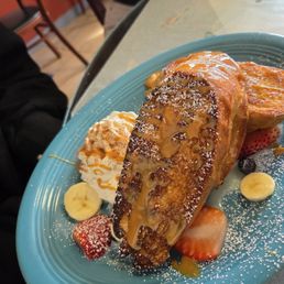 Tres Leches French Toast - A serving of French toast topped with syrup is presented on a blue plate, garnished with sliced strawberries, bananas, and a dollop of whipped cream. The dish is dusted with powdered sugar and appears to be served in a restaurant setting with a marble table surface.