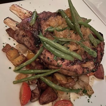 Flatiron Restaurant - Pork chop - A grilled pork chop with visible grill marks is presented on a white plate, topped with a vegetable relish. The plate includes roasted potatoes, cherry tomatoes, and green beans, arranged around the meat. The dish is garnished with chopped herbs, and the presentation emphasizes a hearty, cooked-to-order meal. - Steakhouses Near Me - Brooklyn, New York