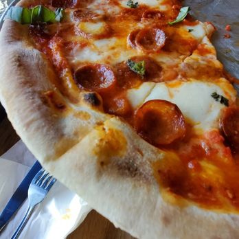 Reno - Reno - A pepperoni pizza with basil leaves and melted cheese on a wooden table, with a person in a blue shirt in the background. - American Near Me - Chicago, Illinois