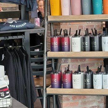 Vibrant Coffeehouse + Kitchen - The tumblers all made by socially responsible company Simple Modern - Multiple rows of tumblers in colors such as yellow, pink, blue, purple, black, and white are displayed on wooden shelves. Each tumbler has a straw, and some feature the 'VIC' logo. The store has a brick wall background, and other retail items like clothing and accessories are visible nearby. - Coffee & Tea Near Me - Moline, Illinois