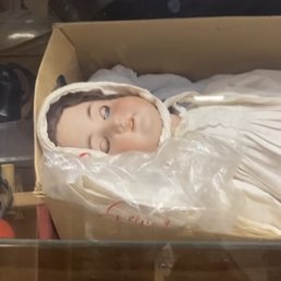 A photo from inside that truly captures my feelings - A doll with detailed facial features, including closed eyes and realistic skin tone, is lying in a cardboard box. The doll is dressed in a white garment with a hood, and it is wrapped in a clear plastic covering. The background shows some indistinct objects, suggesting the doll is stored or displayed in a storage or retail setting.