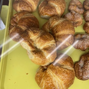 Mr. Donuts - Flaky and buttery delicious croissants - A selection of croissants with a flaky, golden-brown crust are arranged on a yellow tray inside a bakery display case. The croissants vary slightly in shape and size, with some showing layers of pastry. The display case has a glass front, reflecting light and showing part of the surrounding environment. - Donuts Near Me - Oceanside, California