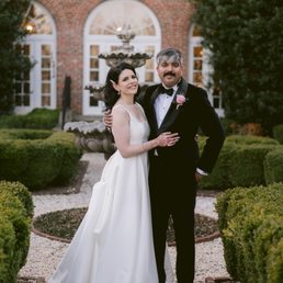 A bride and groom are standing outdoors in a garden area with neatly trimmed bushes and a decorative fountain behind them. The bride wears a white wedding gown with lace details, and the groom is dressed in a black tuxedo with a bow tie and a pink boutonniere. The background features a brick building with large arched windows and a semi-circular window above the fountain. The scene appears to be taken during the evening or late afternoon, with soft lighting.