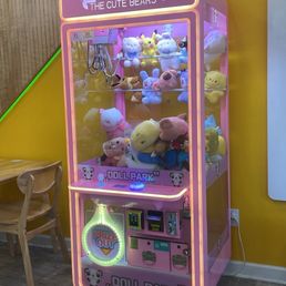 A pink claw machine filled with plush toys, located in a gift shop named 'The Cute Bears,' with a yellow wall and wooden floor.