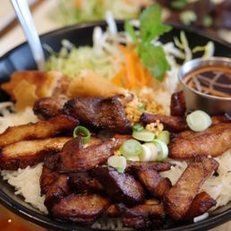 Banh Mi Thit Nuong - A black bowl contains a Vietnamese dish with grilled meats, including chicken and pork, served over rice noodles. The dish is garnished with chopped green onions and fresh herbs, with a side of shredded lettuce, carrots, and cilantro. A small metal cup of dark dipping sauce is included, and other dishes are partially visible in the background.
