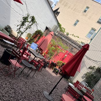 Red Rover - Outdoor patio - The image shows an outdoor patio area with gravel ground, red umbrellas shading metal tables and chairs, and potted plants along the perimeter. String lights are hung across the space, which is enclosed by brick and wooden fences. The background includes a beige building with windows and a clear sky overhead. - Coffee & Tea Near Me - Brooklyn, New York