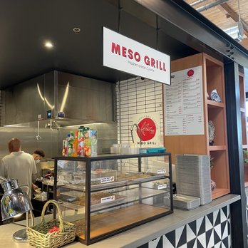 Meso Grill - Inside the Sawmill at Meso Grill - Counter at Meso Grill, a Mediterranean restaurant located inside Sawmill Market, featuring a display case with various food items, a menu on the wall, and staff preparing dishes behind the counter. The decor includes a patterned front panel and wooden shelving with plates. The setting is within an indoor market space with visible ceiling beams and ductwork. - Mediterranean Near Me - Albuquerque, New Mexico