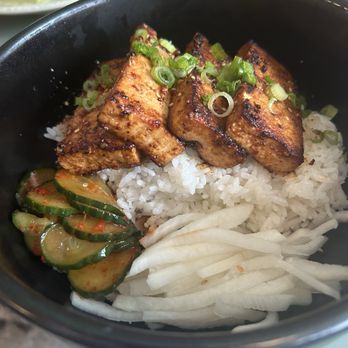 Sonbul - Tofu bowl - A black bowl holds grilled chicken pieces with a caramelized exterior, placed on a bed of white rice. The dish is garnished with chopped green onions. Sliced cucumbers with red pepper flakes and sliced onions are arranged on the side. The background shows a plate of lettuce and another dish, indicating a Korean cuisine meal setting. - Comfort Food Near Me - Brooklyn, New York