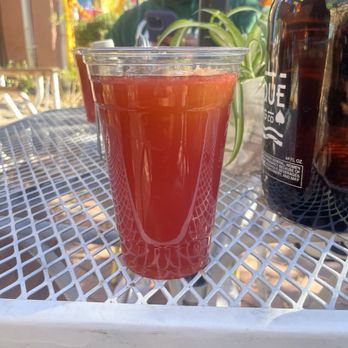 Gobble This - Agua fresca - A clear plastic cup containing a red drink is placed on a white metal outdoor table, with a person taking a photo in the background. - Salvadoran Near Me - Albuquerque, New Mexico