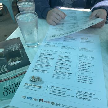 Another Broken Egg Cafe - menu - A person in a blue Harley Davidson shirt is holding a menu at a table with two glasses of water, with chairs and tables visible in the background. - Breakfast & Brunch Near Me - Austin, Texas
