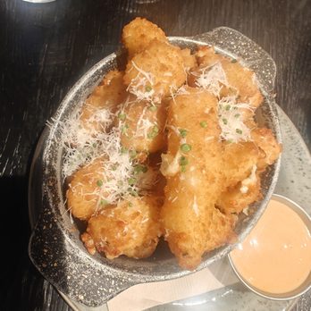 Guinness Open Gate Brewery - Cheese curds - Fried cheese curds are presented on a metal plate, topped with shredded cheese and chopped green onions. The dish is accompanied by a small container of dipping sauce. The plate is placed on a dark wooden table, with drinks visible in the background, indicating a restaurant or bar setting. - Breweries Near Me - Chicago, Illinois