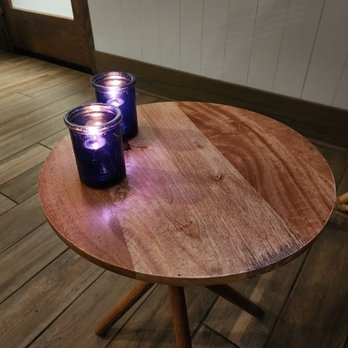 Lindy's Landing - Two purple candles are on a round wooden table with a natural finish, near a door and a wicker chair. - American Near Me - Wauconda, Illinois