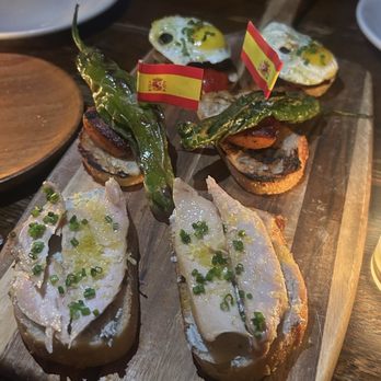 Toro - Pintxos - A wooden platter displays Spanish tapas such as anchovies, peppers, and small fried bites, garnished with small Spanish flags, on a dark restaurant table. - Tapas Bars Near Me - Boston, Massachusetts