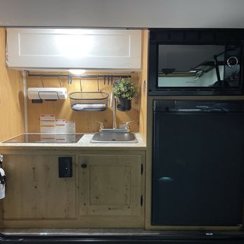 Epic RV Liquidators - The image shows a small RV kitchenette with a microwave oven, a mini refrigerator, a stainless steel sink, and a countertop. The background includes a wooden wall with a mounted paper towel holder, a hanging rack with hooks, and a potted plant. The setup is compact and designed for limited space, typical of recreational vehicle interiors. - RV Dealers Near Me - Madera, California