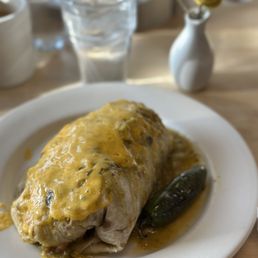 Big Papa Burrito - A burrito topped with melted cheese and sauce is served on a white plate, accompanied by a jalapeño pepper, with a glass of water and a small vase with yellow flowers in the background.