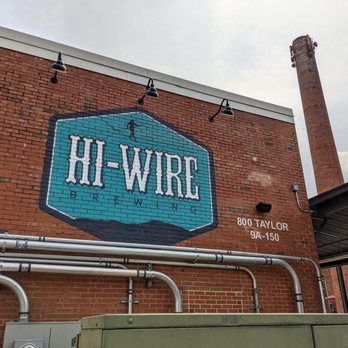 Hi-Wire Brewing - Outside. Hi-Wire Brewing painted on the outside of the brick storefront. Smoke stack in background. - Breweries Near Me - Durham, North Carolina